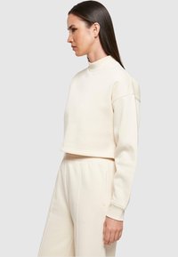 Kreemika värvi lühendatud dressipluus kõrge kaelusega, lõdva istuvusega ja soonikkoes mansettidega, sobitatud kitsenevate pükstega. Sile tekstuur.