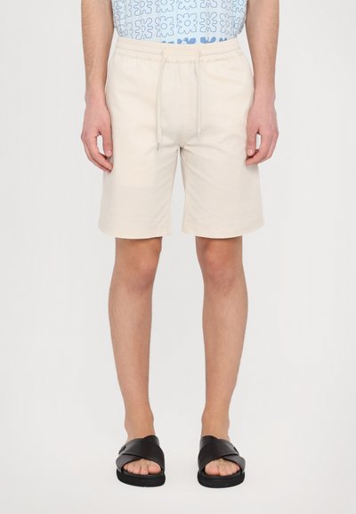 Beige Shorts mit einem elastischen Bund und Kordelzug. Vordertaschen, glatte Textur und knielanger Schnitt. Getragen mit schwarzen Rutsch-Sandalen.