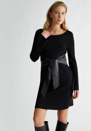Abito di maglia nero con maniche lunghe, caratterizzato da una larga cintura in pelle nera annodata a fiocco in vita. Texture liscia e silhouette aderente.