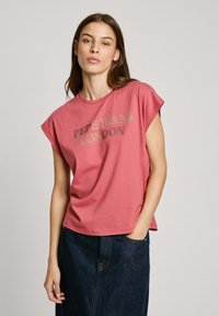 Rožinė medvilninė marškinė su suktomis rankovėmis, ant kurių užrašyta "PEPE JEANS LONDON" metalizuota auksine spalva, derinama su tamsiai mėlynomis džinsinėmis sijoninėmis.