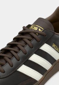 Κοντινό πλάνο καφέ παπουτσιού Adidas Spezial με λευκές ρίγες, καφέ κορδόνια και χρυσό λογότυπο στη γλώσσα και στο πλάι.