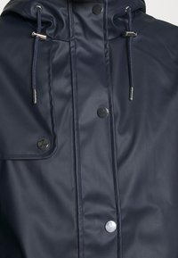 Marinblå vattentät jacka med texturerad yta, har en huva, tryckknappar och justerbara dragsnören. Synlig sömnad och fickor.