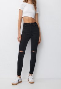 Un jean skinny taille haute noir avec des détails déchirés au niveau des genoux, associé à un t-shirt court blanc et des baskets blanches avec un accent noir.