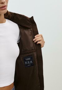 Hnedá zateplená bunda s hladkou vnútornou podšívkou. Obsahuje štítok s nápisom "PADDED COLLECTION" a bočnú kapsu.