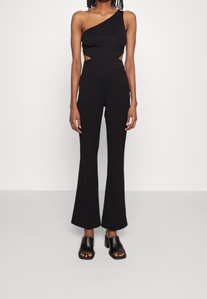 Sort asymmetrisk jumpsuit med udskæringsdetaljer, tætsiddende overdel, vide ben og blød tekstur. Kombineret med sorte åbne sandaler.