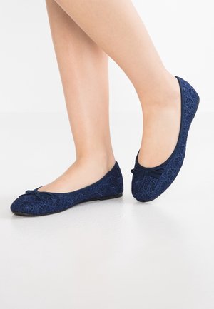 Voeten die marineblauwe ballerina's met linten en kleine strikjes dragen, op een witte ondergrond, één voet iets opgeheven.