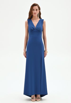 Femme portant une robe de soirée bleue sans manches avec un décolleté plongeant en V et une fente à l'avant, accessoirisée de boucles d'oreilles et bracelets en or, debout droite.