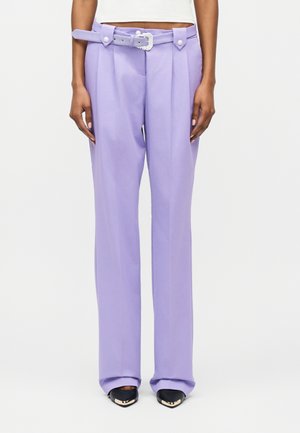 Mujer con pantalones lilas de cintura alta y cinturón a juego, junto con tacones negros de punta afilada, posando frente a un fondo liso.