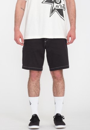 Pantalones cortos negros hechos de un tejido duradero, con costuras blancas, corte recto y bolsillos laterales. Llevados con una camiseta gráfica blanca y zapatos negros.