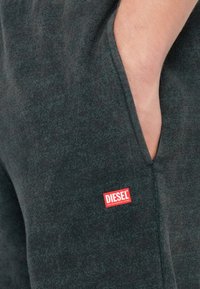 Hand, die in der dunkelgrauen Diesel-Sweatpants-Tasche ruht, mit rotem und weißem Diesel-Logo-Patch nahe der Taschennaht.