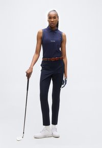 Γυναίκα με μπλε ναυτικό αμάνικο πόλο και παντελόνι κρατάει ρόπαλο του γκολφ, φοράει λευκό γάντι και παπούτσια γκολφ, σε απλό υπόβαθρο.