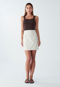 Rjava majica brez rokavov, bež mini krilo z dvema sprednjima žepe, eleganten dizajn, gladka tekstura in črne ravne sandale.