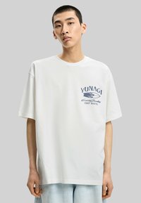 White cotton t-shirt with oversized fit, short sleeves, and graphic print in blue reading “YUNAGI Surf Hostel” on the chest.