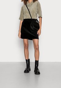 Camicia corta a maniche corte in suede verde oliva, mini gonna in velluto a coste nera, borsa a tracolla rotonda nera con motivo impresso e stivali neri alla caviglia.