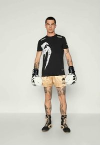 Μαύρη μπλούζα με λευκό σχέδιο, κρέμα σατέν σορτς, μαύρα γάντια πυγμαχίας και μαύρα αθλητικά παπούτσια με μπεζ λεπτομέρειες. Ορατά τα τατουάζ στα πόδια.