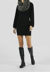 Robe noire en maille à manches longues, assortie à une écharpe infinie en fausse fourrure grise. Le mannequin porte des bottes noires en cuir jusqu'au genou.