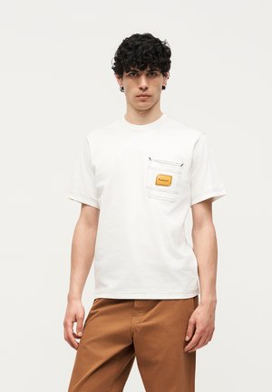 Νέος άνδρας με σγουρά μαλλιά φορώντας ένα λευκό κοντομάνικο πουκάμισο με τσεπάκι στο στήθος και καφέ παντελόνι, στέκεται μπροστά σε απλό φόντο.