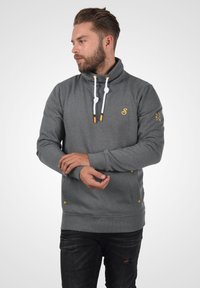 Sudadera gris con un alto cuello, cordones blancos y acentos amarillos. Cuenta con dos bolsillos laterales y puños acanalados. Diseño simple y funcional.
