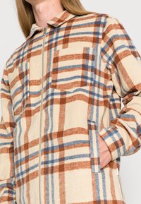 Kobieta w beżowej, brązowej i niebieskiej kraciastej kurtce zapinanej na zamek z kieszonką na piersi, stojąca z prawą ręką w kieszeni bocznej.