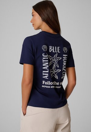 Kobieta z długimi ciemnymi włosami, ubrana w granatowy t-shirt z białymi wzorami rozgwiazd i muszli oraz napisem na plecach, i jasnobeżowe spodnie.