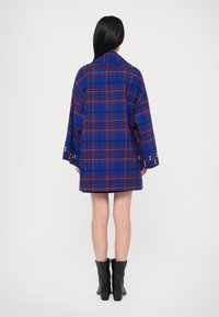 Casaco xadrez azul com detalhes em vermelho e preto, ajuste oversized, mangas largas e detalhes em botão metálico, usado sobre uma saia preta e botas.