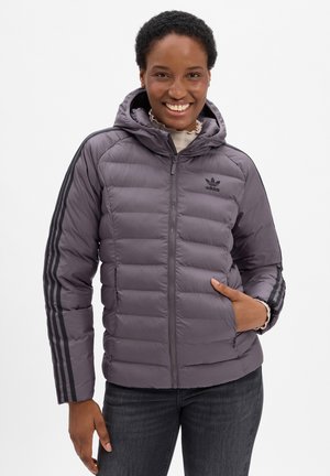 Uśmiechnięta kobieta w szarej puchowej kurtce Adidas z kapturem i czarnymi paskami na rękawach, pozująca z jedną ręką w kieszeni.