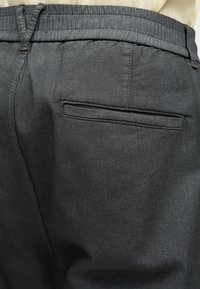 Pantalon gris foncé avec taille élastique et poche arrière passepoilée simple, porté avec un haut de couleur claire.