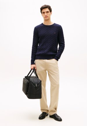 Marineblaue Strickjacke mit Zopfmuster, beige Hose, schwarze Leder Tasche mit zwei oberen Griffen, glatte Textur, dezente Metallakzente, lässiges Outfit.