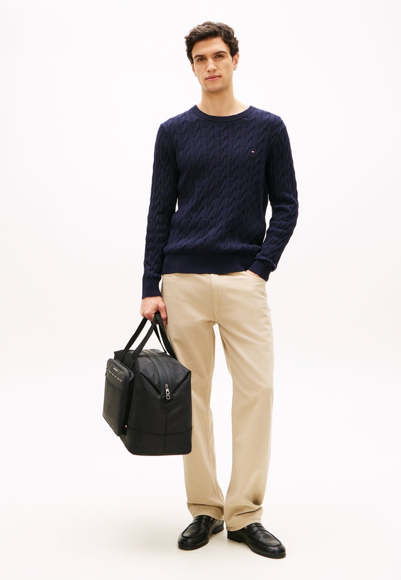 Μπλε σκούρο πουλόβερ με πλέξη καλώδιο, μπεζ παντελόνι, μαύρη δερμάτινη τσάντα με δύο πάνω λαβές, λεία υφή, διακριτική λεπτομέρεια εξαρτημάτων, καθημερινή εμφάνιση.