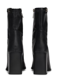 Botas de tobillo de cuero negro con una textura suave, tacones altos en bloque y cremalleras laterales. Presentan detalles de hardware dorado en la parte superior.