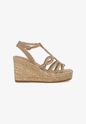 Sandale compensée avec une plateforme en jute tressé et un dessus en suède beige. Présente des brides décoratives avec des gemmes transparentes. Bride de cheville réglable.