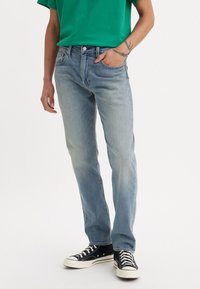Vaqueros azules descoloridos con corte recto, tejido de mezclilla, diseño de cinco bolsillos y bordes desgastados, combinados con zapatillas de lona negras.