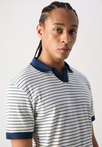 Polo shirt blanc à rayures avec col et accents de manches bleu marine. Texture côtelée, design sans boutons et manches courtes.