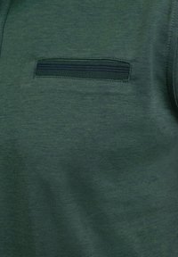 Tejido verde texturizado con un bolsillo horizontal con cremallera. La superficie posee un patrón sutil, ofreciendo una apariencia suave.