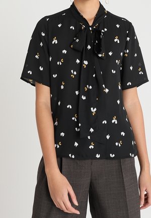 Une personne porte un chemisier noir à manches courtes avec un col à nœud et un petit imprimé floral blanc et jaune, associé à un pantalon gris à carreaux.