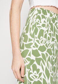 Pantalones de patrón floral verde con fondo blanco, que cuentan con una cinturilla elástica y un corte relajado. Textura de tela suave visible.