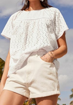 Femme avec la main dans la poche, portant un chemisier en dentelle blanche à œillets et un short blanc retroussé, en plein air sous un ciel bleu.