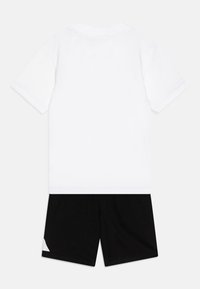Camiseta blanca de algodón con mangas cortas, combinada con pantalones cortos negros que tienen un acento blanco en la parte inferior izquierda. Diseño simple y minimalista.