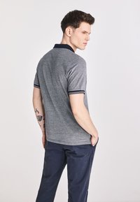 Szara koszulka polo z granatowym kołnierzykiem i paskowanymi mankietami, wykonana z miękkiego materiału, w zestawieniu z granatowymi spodniami. Widoczne tatuaże na ramieniu.