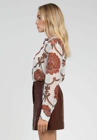 Blouse à manches longues en tissu blanc avec des motifs floraux bruns, associée à une jupe en cuir marron ornée de clous en argent.