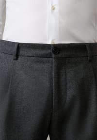 Pantaloni grigi texture con taglio dritto, dotati di chiusura frontale con bottone e tasche laterali, abbinati a una camicia bianca.