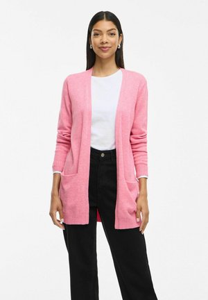 Cardigan en tricot rose avec des manches longues et une ouverture devant, comportant deux poches avant et des poignets côtelés. Porté sur un haut blanc et un pantalon noir.