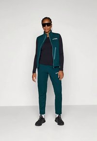 Kvinne iført teal ermeløs vest og bukser med svart langermet skjorte, svarte sneakers, hvite sokker og store svarte solbriller som står mot en hvit vegg.