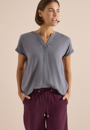 Kobieta z krótkimi brązowymi włosami nosi luźną szarą bluzkę z dekoltem w serek i bordowe spodnie z troczkiem, ręce w kieszeniach, patrzy w prawo na beżowym tle.