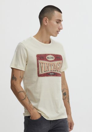 Cremefarvet t-shirt lavet af blødt bomuldsstof, med et slidt rødt og sort vintage-inspireret grafisk tryk på fronten. Korte ærmer og rund halsudskæring.