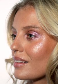 Nahaufnahme eines Gesichts mit schimmerndem rosa Lidschatten, glänzender Haut und glänzenden Lippen. Helles, blondes Haar umrahmt teilweise die Gesichtszüge.