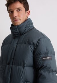 Ciemnoturkusowa pikowana kurtka puchowa z wysokim kołnierzem, zapięciem na zatrzaski, pikowanym wzorem oraz naszywką z logo na lewym ramieniu.