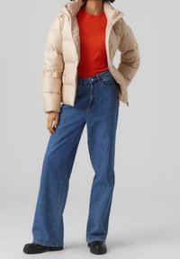 Beige puffer-takki kiiltävällä pinnalla, punainen istuva paita ja siniset leveälahkeiset farkut. Mustat saappaat täydentävät asun.