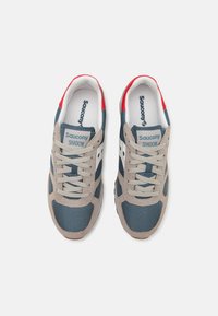 Saucony Shadow sneakers har en grå og marineblå mesh-overdel med beige semsket skinn-detaljer, hvit mellomsåle og rød hæltab. Klassisk snøring.