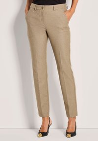 Vrouw in beige slim-fit geblokte broek met zakken en zwarte hoge hakken met gouden tenen, staand met handen in de zakken.